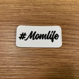 May include: A white rectangular patch with rounded corners. The patch features the black text "#Momlife" in a cursive font. The patch is likely an iron-on or sew-on patch for clothing or accessories.