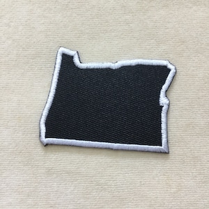 May include: A black embroidered patch in the shape of the state of Oregon. The patch has a white stitched border and is set against a neutral background. The patch is likely for clothing or accessories.