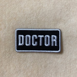 May include: A rectangular black and white embroidered patch with the word "DOCTOR" in white capital letters. The patch has a black background and a white border. The patch is on a beige surface.