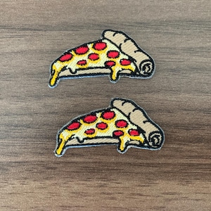 May include: Two embroidered patches shaped like pizza slices. Each slice features a tan crust, yellow melted cheese, and red pepperoni circles. The embroidery has a black outline and depicts the cheese dripping down the side of the pizza slice.