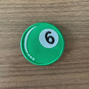 May include: A round, green embroidered patch featuring a white circle with the number 6 in black. The patch has a white curved line and three white dots, resembling a billiard ball. The patch is on a wooden surface.