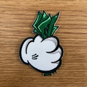 May include: Embroidered patch featuring a white cartoon hand holding a bundle of green money. The hand has black outlines and three black lines on the palm. The money has green and white details. The patch is on a wooden surface.