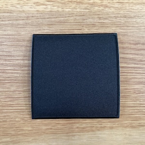 May include: A square, navy blue patch with a black border. The patch has a textured surface and is set against a light brown, wood-grain background. The patch is a solid color with no visible design.