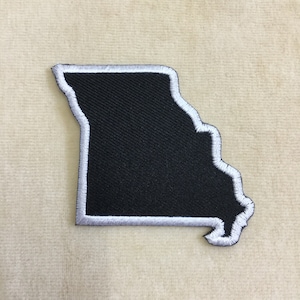 May include: A black patch in the shape of the state of Missouri, with a white embroidered border. The patch is made of a textured fabric and is likely designed to be ironed or sewn onto clothing or accessories.