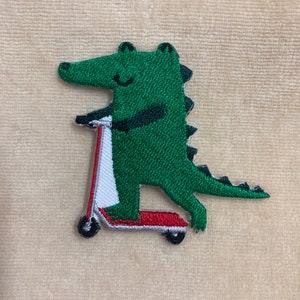 May include: Embroidered patch featuring a green alligator riding a red, white, and black scooter. The alligator has a smiling face and is wearing a black shirt. The patch is likely for clothing or accessories.