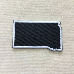 May include: A black patch in the shape of the state of South Dakota, with a white embroidered border. The patch is rectangular with a jagged edge on the eastern side. The patch is likely for clothing or accessories.
