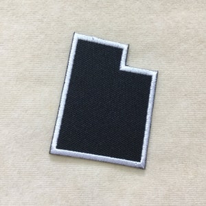 May include: A black embroidered patch in the shape of the state of Utah, with a white border. The patch is made of a textured fabric and is set against a light beige background.