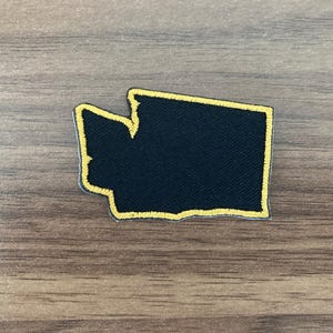 May include: A black embroidered patch in the shape of the state of Washington. The patch features a solid black center with a thick, bright yellow stitched border that outlines the entire geographic silhouette of the state.