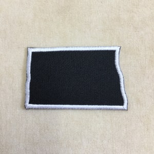 May include: A rectangular black patch with a white border. The patch has a slightly irregular edge on one side, giving it a unique shape. The white border is created with a stitched design.