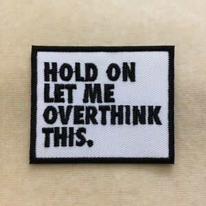 May include: A rectangular white patch with black embroidered text that reads "HOLD ON LET ME OVERTHINK THIS." The patch has a black border and is likely an iron-on or sew-on patch for clothing or accessories.