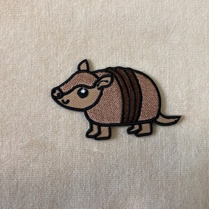 May include: Embroidered patch featuring a cartoon armadillo. The patch has a tan body with dark brown stripes and a black outline. The armadillo has a smiling face with a white eye and a small black eye. The patch is on a beige background.