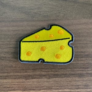 May include: A yellow cheese slice patch with embroidered orange holes and a black outline. The patch is on a wooden surface. This patch is a fun accessory for bags, jackets, or other fabric items.