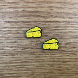 May include: Two yellow cheese wedge patches with black embroidered outlines and details. The patches are on a wooden surface. The cheese wedges have a textured appearance, suggesting a fabric or embroidered design.