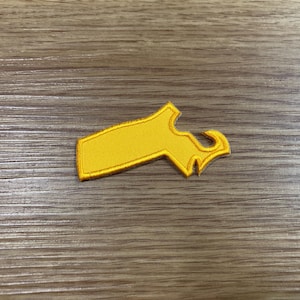 May include: A yellow embroidered patch in the shape of a stylized key. The patch has a rectangular body with a decorative top and a curved, hook-like element. The patch is on a wooden surface.