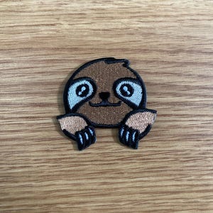 May include: Embroidered patch featuring a smiling sloth with brown fur, light blue eyes, and tan paws. The sloth is outlined in black and appears to be peeking out. This patch is suitable for clothing, bags, or other fabric items.