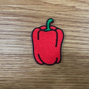 May include: A red bell pepper patch with a green stem and black outline. The patch is embroidered with a textured appearance. The patch is on a wooden surface.