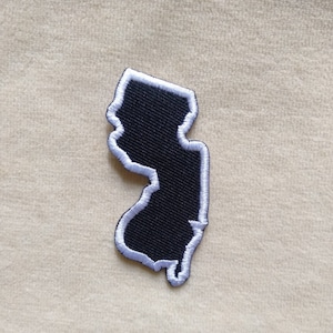 May include: Embroidered patch in the shape of New Jersey. The patch features a black interior with a white outline and border. The patch is likely designed to be ironed on or sewn onto fabric.