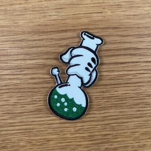 May include: An embroidered patch featuring a cartoon-style design of a laboratory flask with a hand holding it. The flask is light blue with a green liquid and white dots. The hand is white with black details.