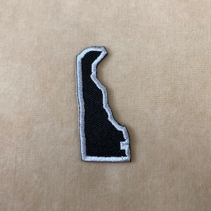 May include: A black and white embroidered patch in the shape of the state of Delaware. The patch has a black interior and a white border. The patch is on a tan fabric surface.