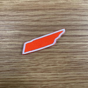 May include: An orange patch in the shape of the state of Tennessee, outlined in white. The patch is on a wooden surface.