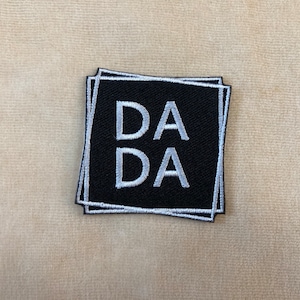 May include: Black embroidered patch with the word "DADA" in white, stacked on top of each other. The patch has a white border and is set against a beige background. The patch is square-shaped.
