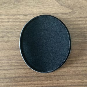 May include: A circular black patch with a smooth, fabric-like texture and a clean, stitched border. The patch is solid black throughout, featuring a simple, uniform design with a defined edge that separates the center from the outer perimeter.