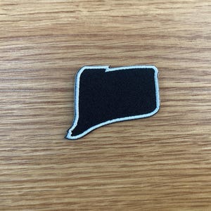 May include: A black patch in the shape of a speech bubble, outlined in white. The patch is made of a textured fabric and is set against a wooden background. This patch could be used to decorate clothing or accessories.