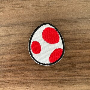 May include: An embroidered patch in the shape of an oval egg. The patch features a white background with three distinct red spots of varying sizes. The entire edge of the egg-shaped patch is outlined with a thick black embroidered border.