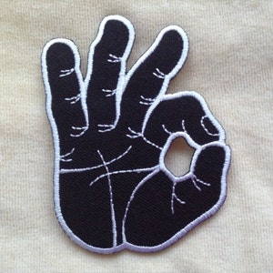 May include: Black embroidered patch in the shape of a hand making the "OK" gesture. The patch has a black background with white stitching outlining the hand and fingers. The palm lines are also stitched in white.