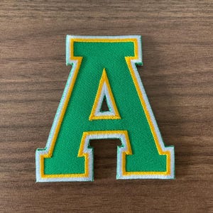 May include: A varsity-style letter A patch featuring a green center with a yellow inner border and a white outer border. The embroidered patch has a classic block font design with clean, stitched edges, suitable for application onto various fabric items.