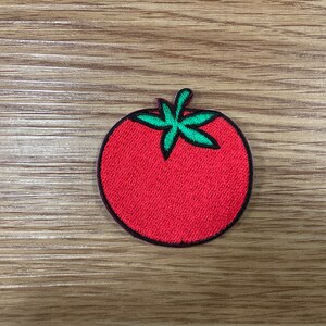 May include: A circular embroidered patch featuring a red tomato with green leaves. The patch has a black border and is set against a wooden background. This patch is suitable for clothing or accessories.