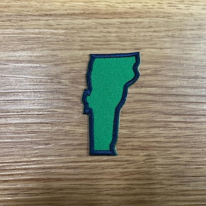 May include: A green patch in the shape of the state of Vermont, with a dark blue border. The patch is on a wooden surface.