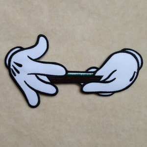 May include: Embroidered patch featuring white cartoon hands with black outlines, holding a brown object with a green stripe. The patch is set against a tan background. The hands are in a gesture of rolling.