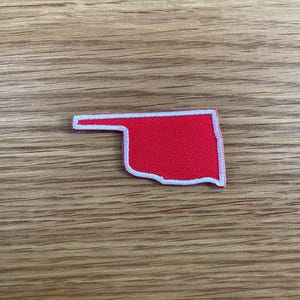 May include: A red patch in the shape of the state of Oklahoma, with a white border. The patch is made of fabric and is set against a wooden background.