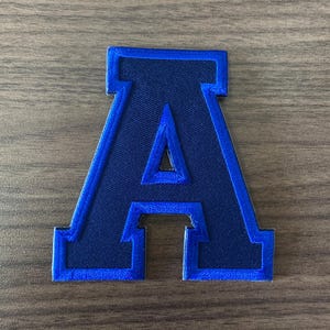 May include: A navy blue embroidered letter "A" patch with a bright blue border. The patch is a stylized varsity letter, perfect for customizing apparel or accessories. The patch is on a wooden surface.