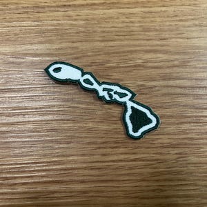 May include: A white and dark green embroidered patch in the shape of the Hawaiian Islands. The patch is on a wooden surface. The patch has a white outline and a dark green interior.