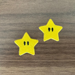 May include: Two yellow star-shaped embroidered patches. Each five-pointed star features a pair of vertical black embroidered lines in the center, resembling eyes. The patches have a textured fabric surface and a distinct white border edge.