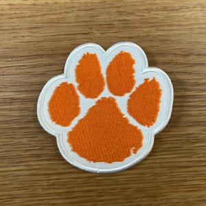 May include: Embroidered patch in the shape of a paw print. The paw print is orange with a white border. The patch is likely for clothing or accessories, and is a symbol of a pet or animal lover.