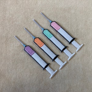 May include: Four embroidered syringe patches with colorful tops in purple, orange, green, and pink. Each patch has a white body, black accents, and a silver needle. The patches are arranged diagonally on a beige surface.