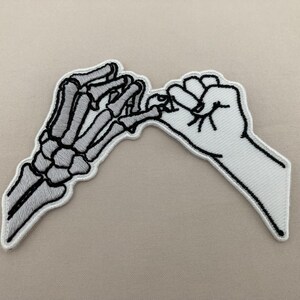 May include: Embroidered patch featuring two hands in a pinky promise gesture. One hand is a skeletal design in gray and white, while the other is a white hand with black outlines. The patch has a white border and is set against a neutral background.