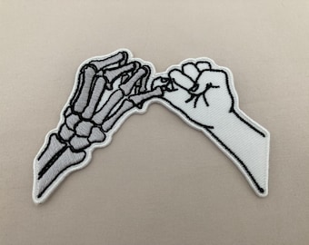 May include: Embroidered patch featuring two hands in a pinky promise gesture. One hand is a skeletal design in gray and white, while the other is a white hand with black outlines. The patch has a white border and is set against a neutral background.