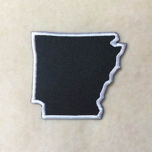 May include: A black embroidered patch in the shape of the state of Arkansas. The patch has a white border and is made of a textured fabric. The patch is likely designed to be ironed or sewn onto clothing or accessories.