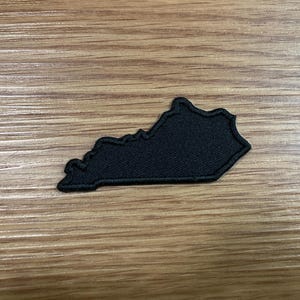 May include: A solid black embroidered patch in the shape of the state of Kentucky. The patch features a detailed outline of the state borders with a dense, textured fabric construction and a matching black stitched border around the entire edge.