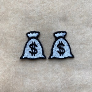 May include: Two embroidered patches shaped like money bags. Each patch is white with a black outline and a black dollar sign. The patches are on a beige fabric background.