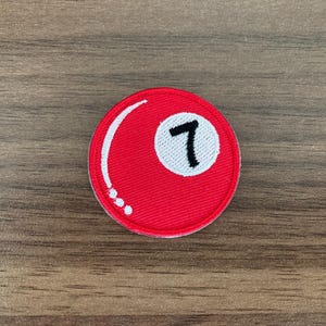 May include: A circular embroidered patch featuring a vibrant red background with a white circular inset containing the black number seven. The design mimics a billiard ball, complete with a white curved line and three small white dots.