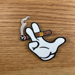 May include: Embroidered patch featuring a white cartoon hand making a peace sign, holding a brown cigar with a yellow band. The cigar has smoke rising from it. The patch has a black border.