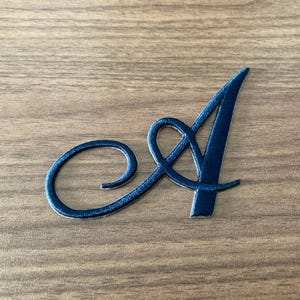 May include: A dark navy blue letter A in a cursive script font. The letter features a smooth, matte finish with a distinct, flowing tail that loops around the left side of the character, creating a decorative and elegant appearance for the alphabet.