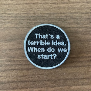 May include: Round black patch with white text that reads "That's a terrible idea. When do we start?" The patch has a white border and is set against a wooden background.