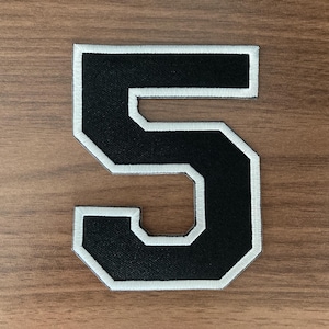 May include: A black embroidered patch in the shape of the number five, featuring a bold block font style with a clean white border outline. The patch has a textured fabric surface and is designed for application onto various textile items.
