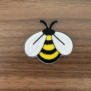 May include: An embroidered patch in the shape of a bee. The bee features a black and yellow striped abdomen, a black head with two black antennae, and two large white wings with black outlines. The design is flat and has a visible stitched texture.